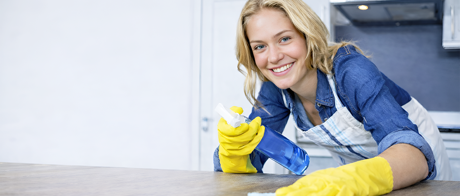 A smiling woman wearing a blue shirt and yellow cleaning gloves, wiping a surface with a cloth and holding a spray bottle, in a kitchen setting with a blurred background.