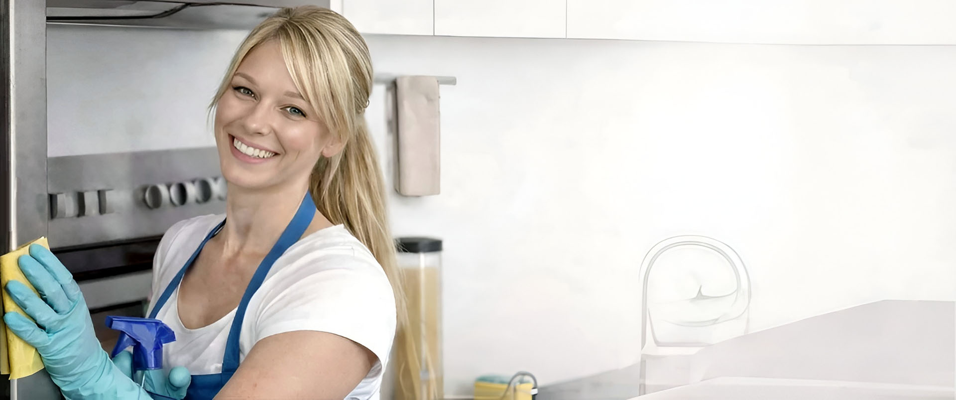 A smiling woman wearing a white top and blue apron, cleaning a kitchen appliance with yellow gloves, standing in a modern kitchen with a stove and sink in the background.