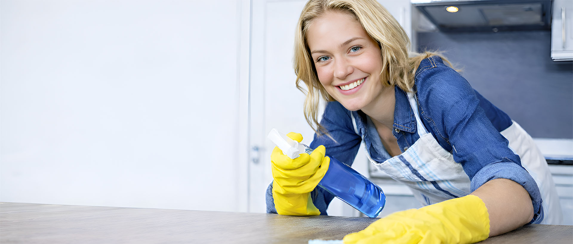 A smiling woman wearing a blue shirt and yellow cleaning gloves, wiping a surface with a cloth and holding a spray bottle, in a kitchen setting with a blurred background.